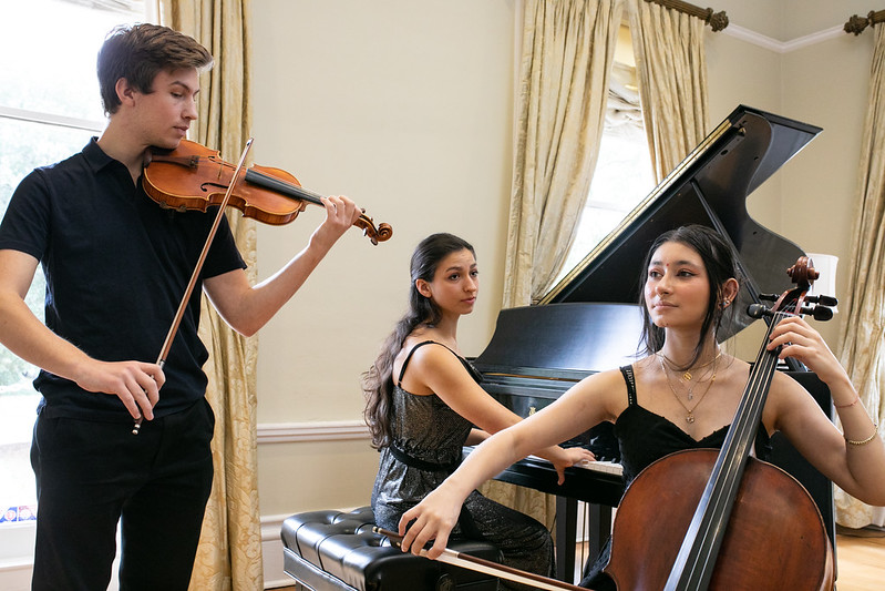 One student standing playing the violin, one seated playing the cello, one playing the piano