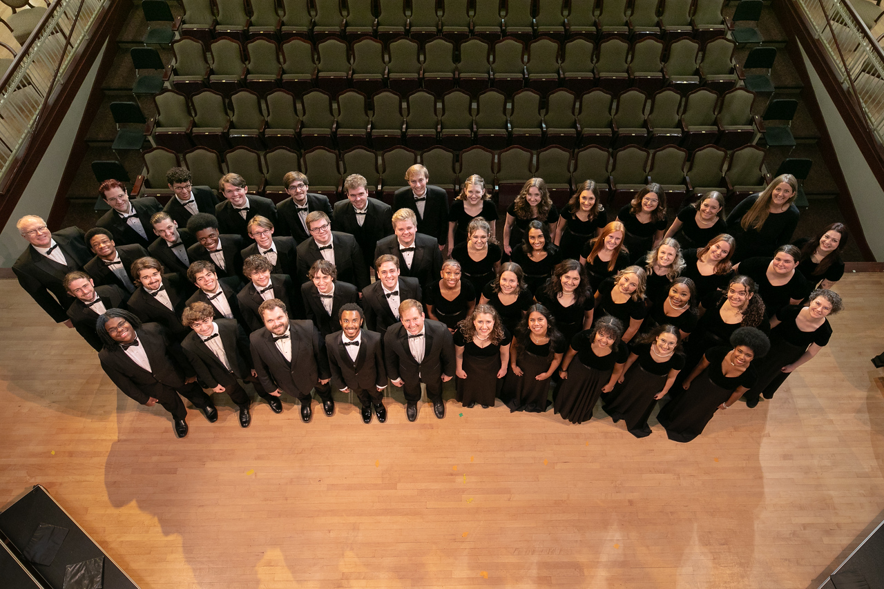 Four rows of singers all in black, standing on stage