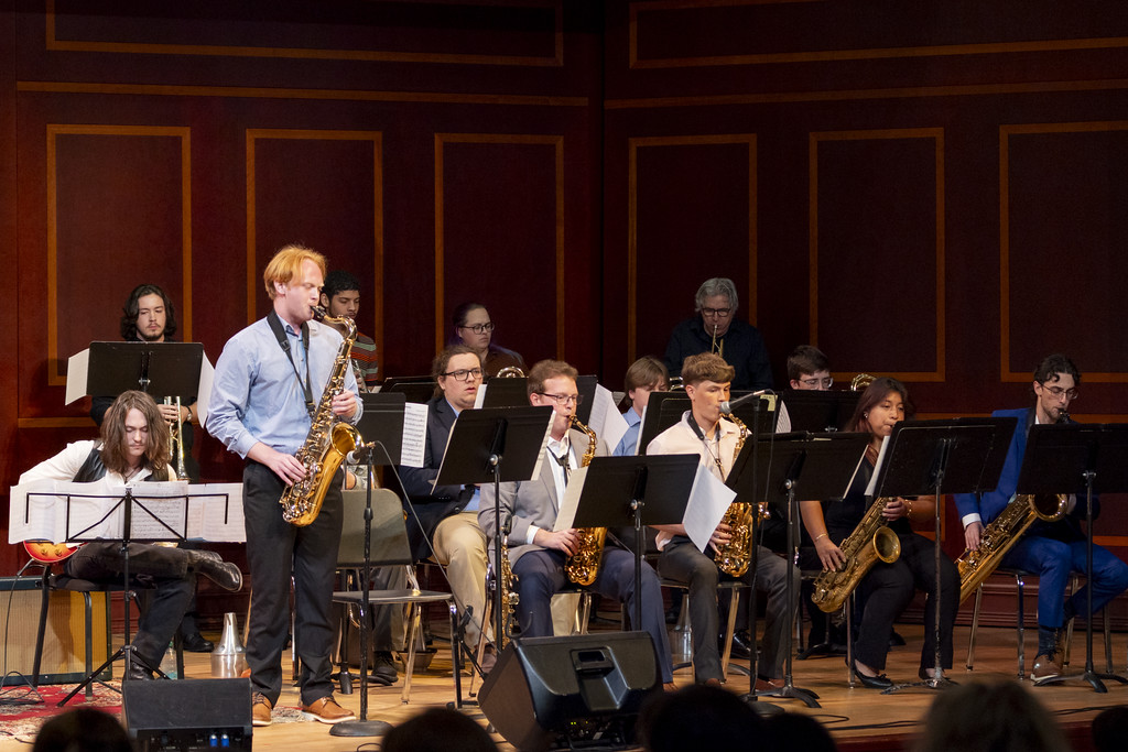 jazz ensemble players sit on stage, saxophone soloist stands