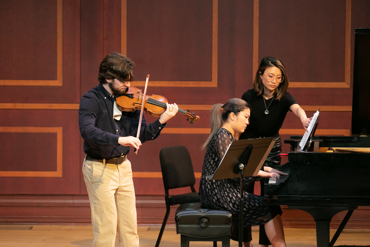 A violinist stands and a piano player is at the piano along with a page-turner